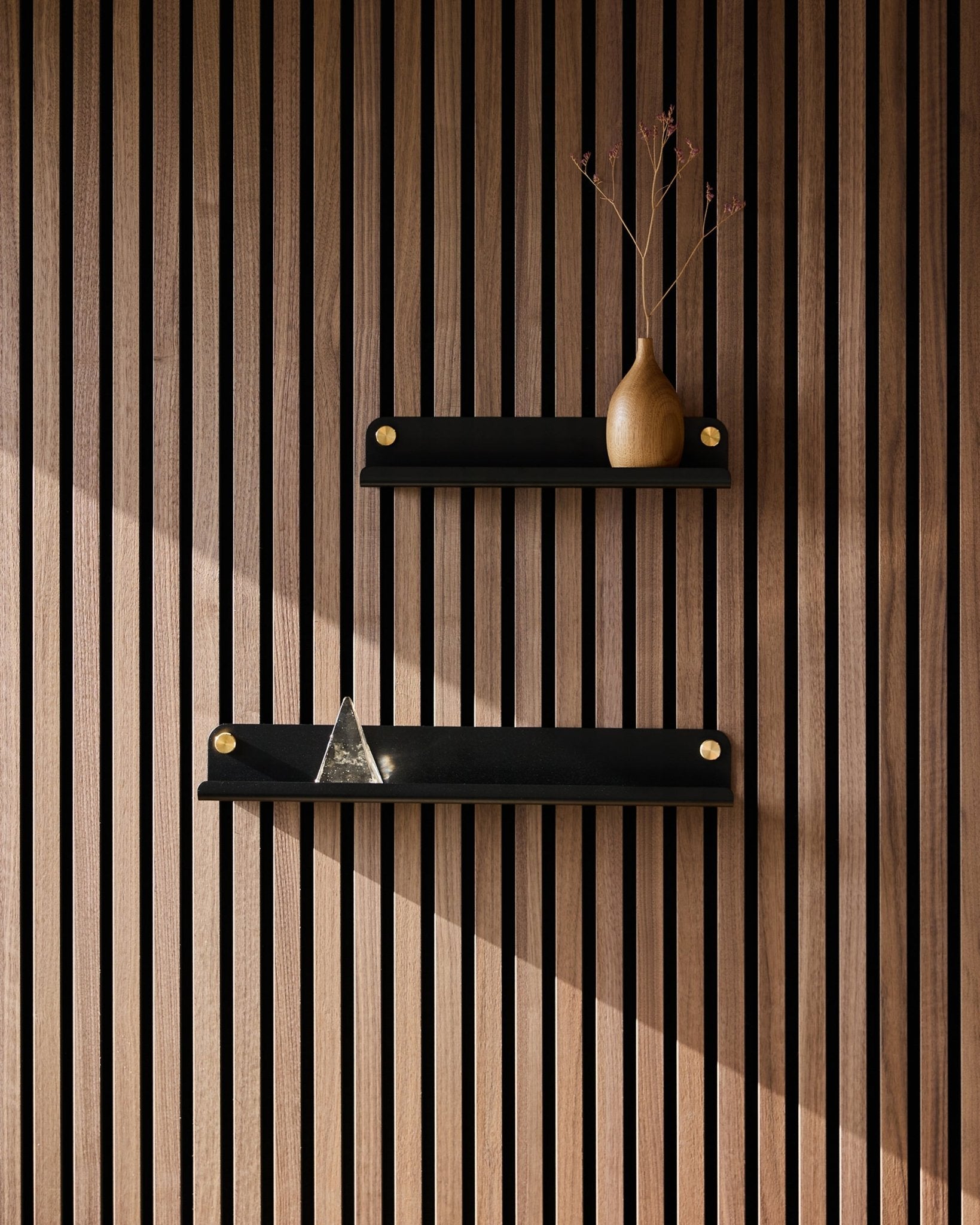 Dos estantes estrechos WoodUpp Norr black (35,9 cm) montados en un panel acústico de lamas de estilo nórdico; el estante superior con un jarrón de ramas secas, el inferior con una pirámide de cristal.